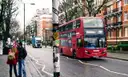 A red double-decker bus on Abbey Road, with two people waiting at the kerb