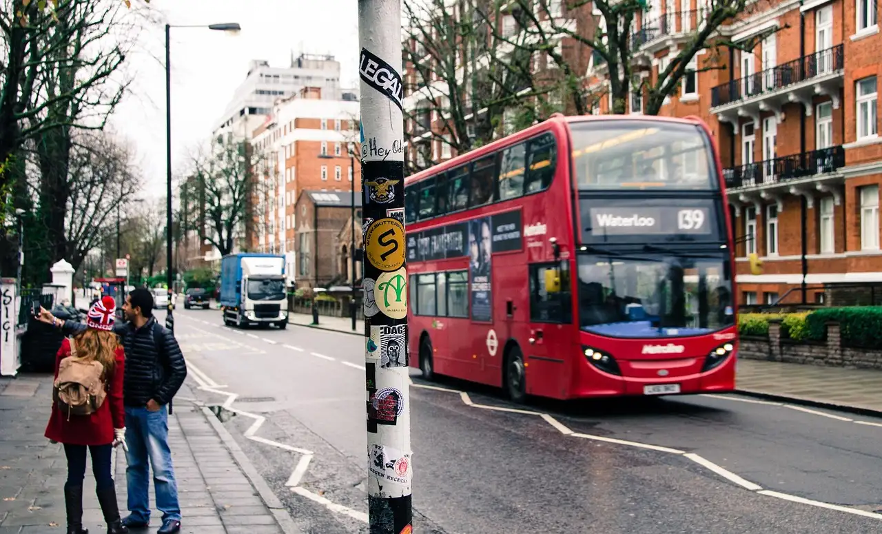 A red double-decker bus on Abbey Road, with two people waiting at the kerb