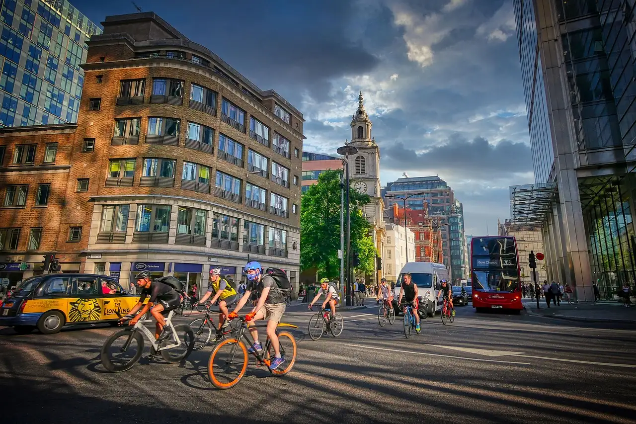A group of cyclists riding down a sunny City of London street, a red bus behind them