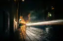 A dark cobbled street at night with a lamp glowing and light trails