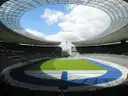 An empty stadium seen from high in the stands, with seats around the pitch under a blue sky