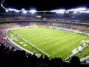 A floodlit football match with fans watching from the stands
