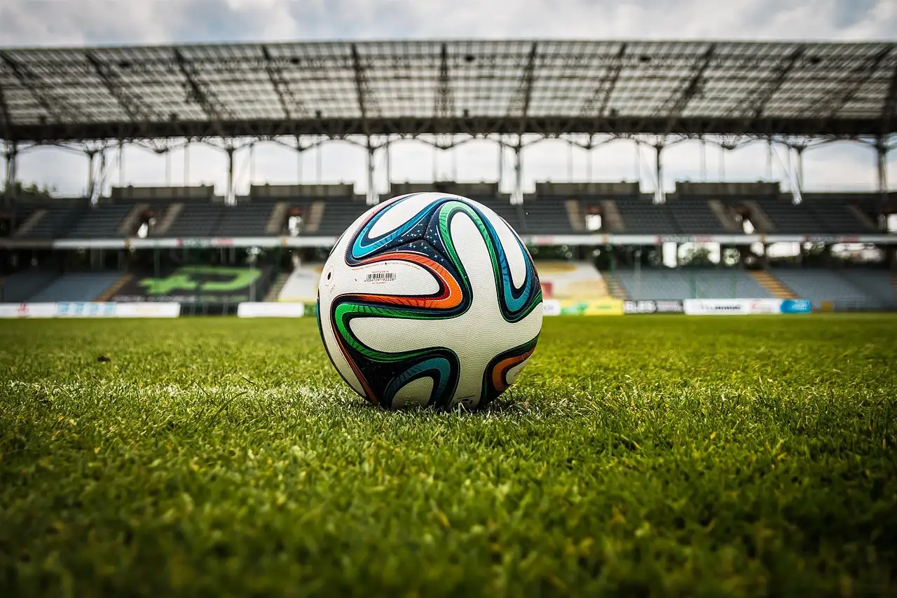 A football on the grass of a stadium pitch with the stands behind