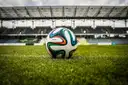 A football on the grass of a stadium pitch with the stands behind