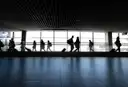 Travellers with luggage in silhouette walking along a bright airport terminal window