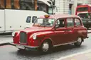 A red London taxi with a classic rounded front driving along a city street, buses behind