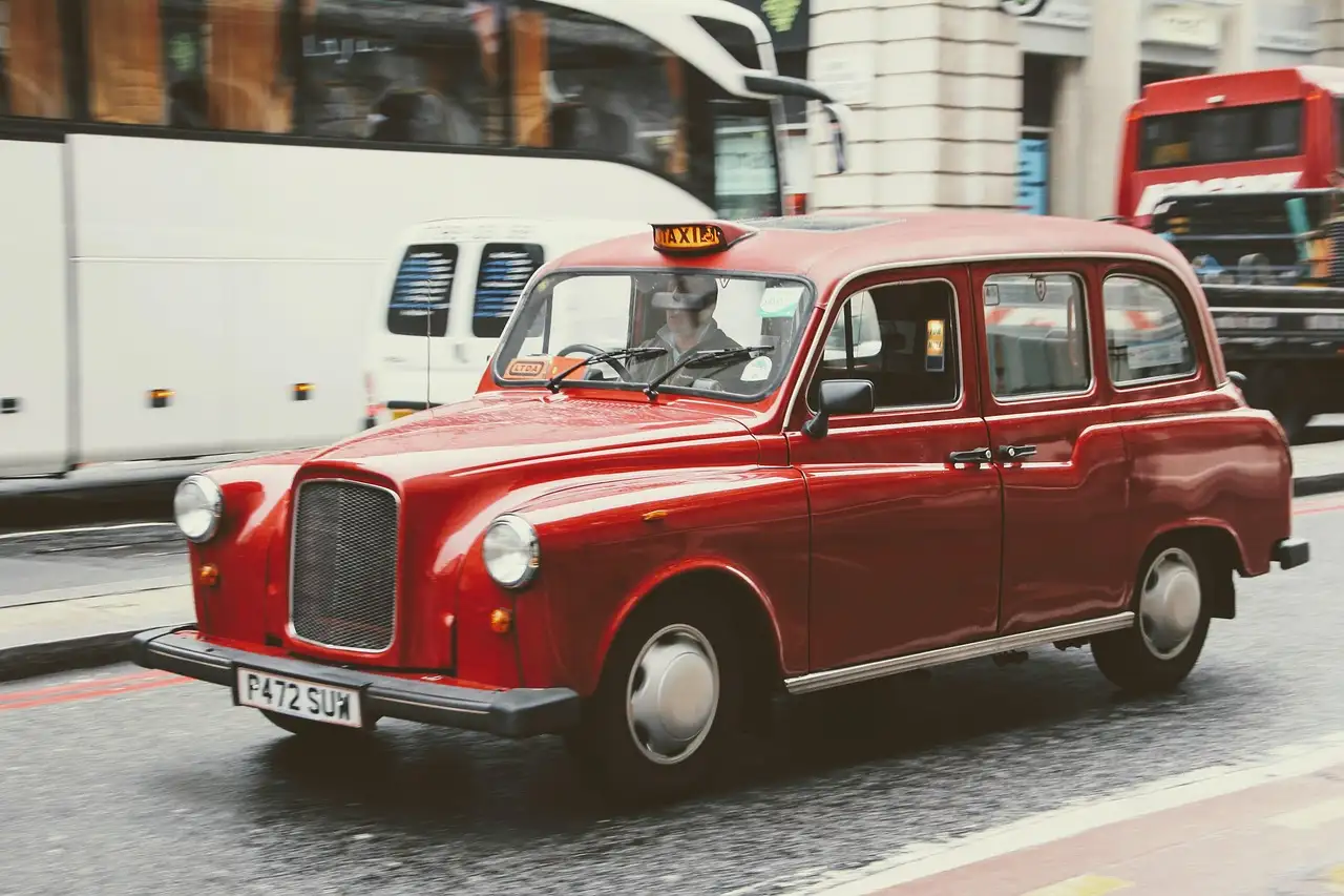 A red London taxi with a classic rounded front driving along a city street, buses behind