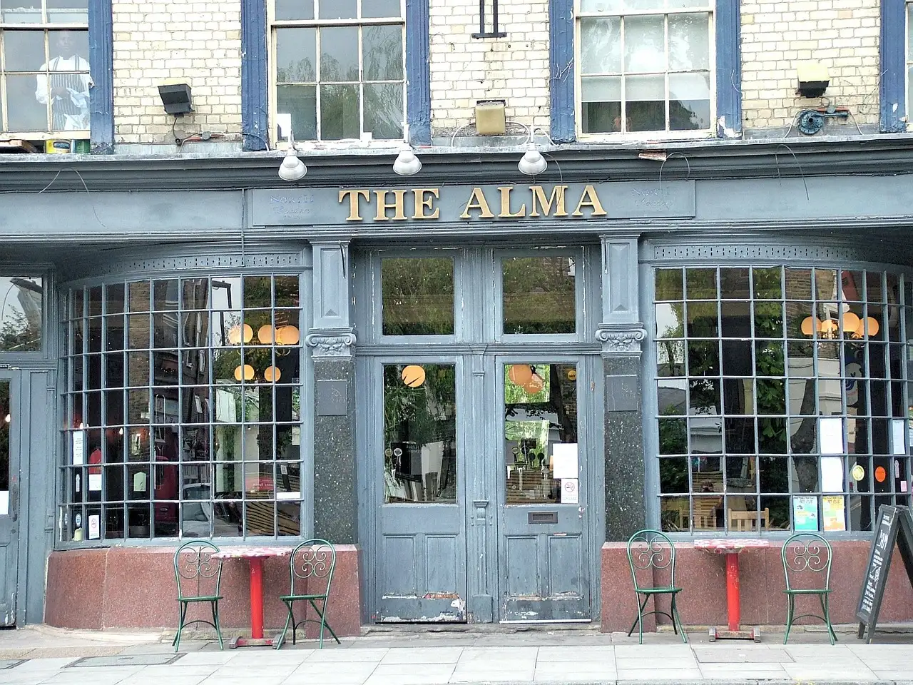 The blue-grey front of a London pub with tall small-paned windows and two café tables outside