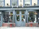 The blue-grey front of a London pub with tall small-paned windows and two café tables outside