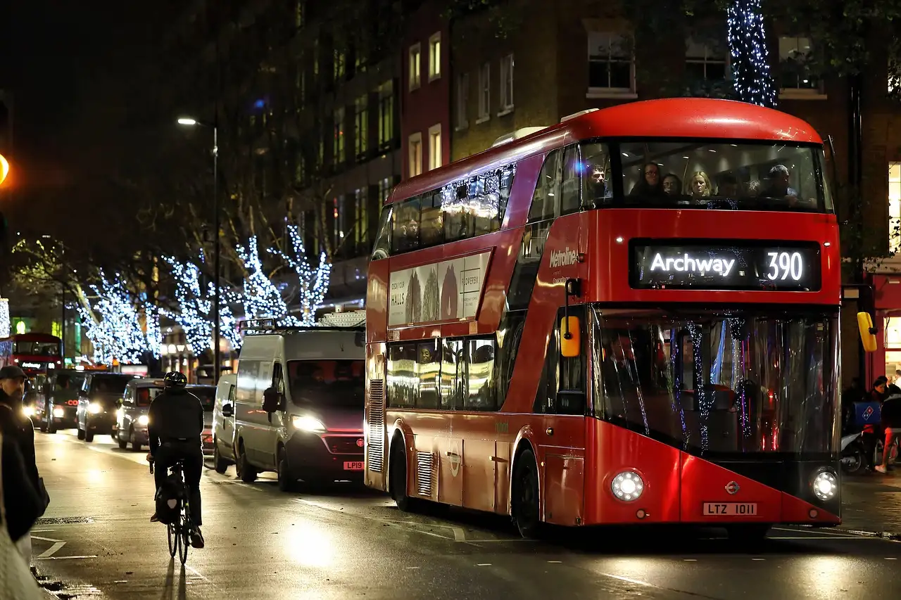 A red double-decker bus on a London street at night with Christmas lights in the trees