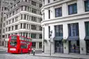 A red double-decker bus turning a corner between London office buildings