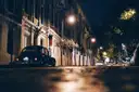 A dark street at night lit by street lamps, with an old car parked by tall houses