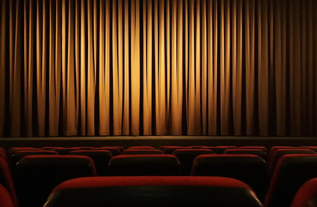 A lit stage curtain behind rows of empty red theatre seats