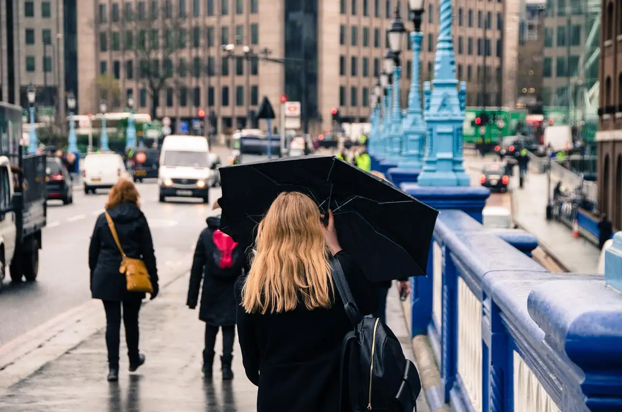 People walking over a London bridge with blue railings in the rain, one woman holding a black umbrella
