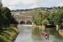 Pulteney Bridge arching over the River Avon in Bath with a red narrowboat on the water and the town rising on the hill behind