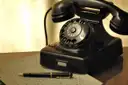 A black rotary telephone on a desk next to a handwritten letter and fountain pen